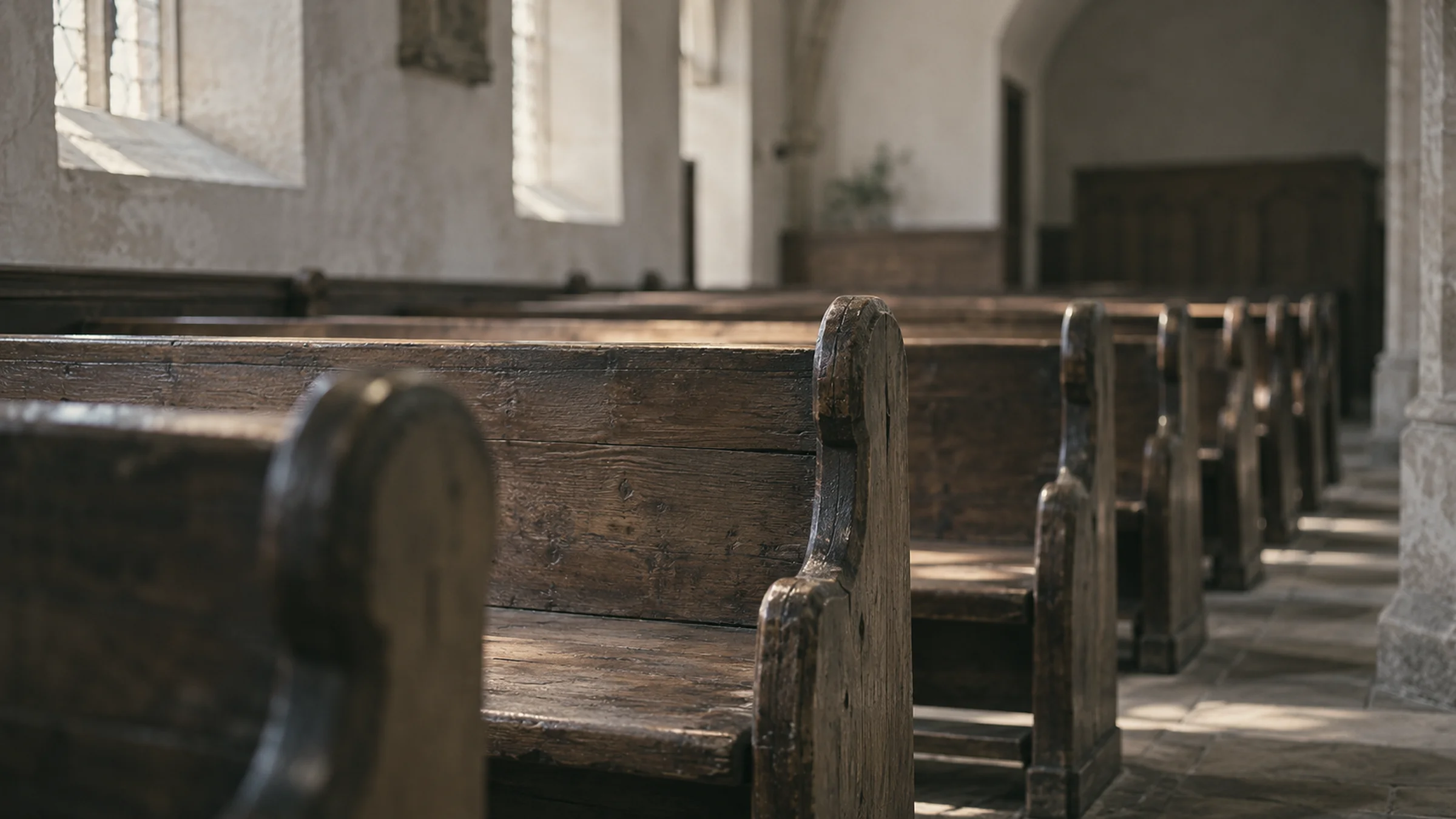 Rows of empty wooden church pews in side light, with plaster walls and no people in the frame.